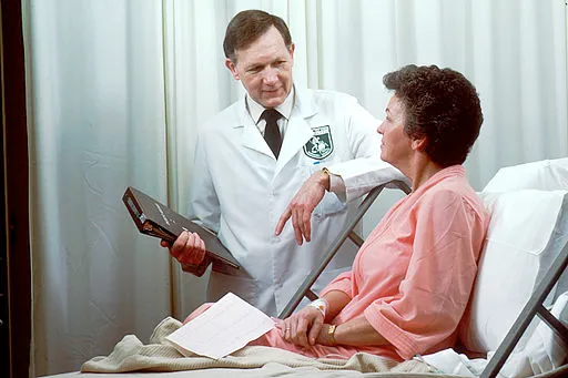 A Caucasian male doctor from the Oncology Branch consults with a Caucasian female adult patient, who is sitting up in a hospital bed